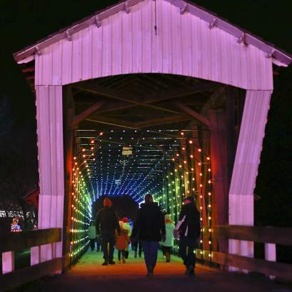People walking through a wooden covered bridge at night, with colorful lights lining the interior and creating a glowing tunnel effect.