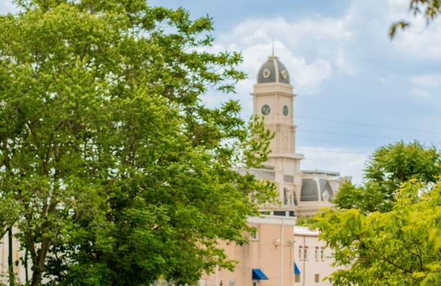 Historic courthouse building surrounded by trees and greenery, photographed in warm daylight. People ride rail bikes in the foreground.