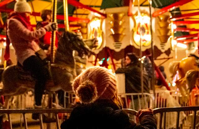People seated on painted carousel horses riding a brightly lit carousel at night, with warm yellow and red lights and decorative panels visible around the ride.
