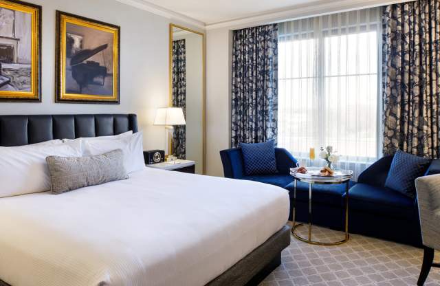 A hotel room with a king-size bed dressed in white bedding, a dark upholstered headboard, and framed artwork above the bed. The room includes a seating area with blue chairs, a small table, floor lamps, and patterned curtains framing a large window, creating a modern, upscale atmosphere.