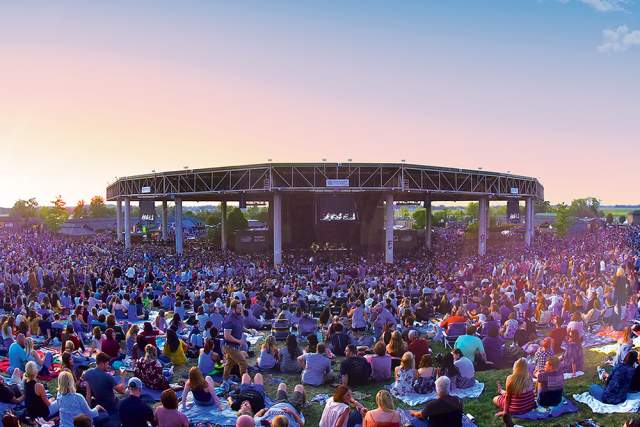 Large outdoor crowd seated on blankets and lawn chairs facing the stage at Ruoff Music Center during a sunset concert, with the amphitheater roof centered in the background.