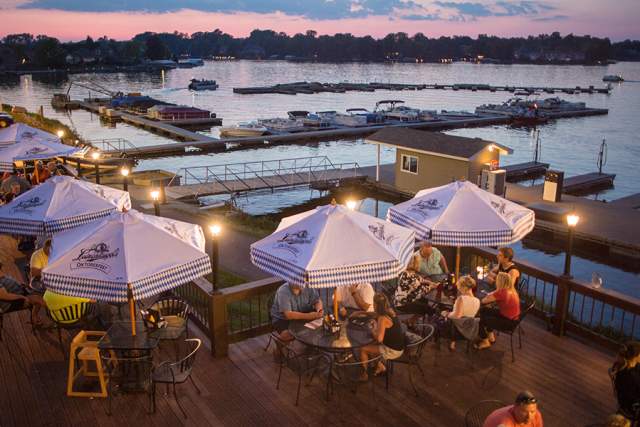 An outdoor patio with tables, diners, and umbrellas overlooking a body of water during early evening.