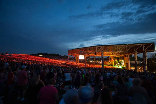 Large outdoor concert venue at dusk, with a brightly lit stage and a crowd filling the seating area. Rows of lights glow across the venue under a darkening blue sky.