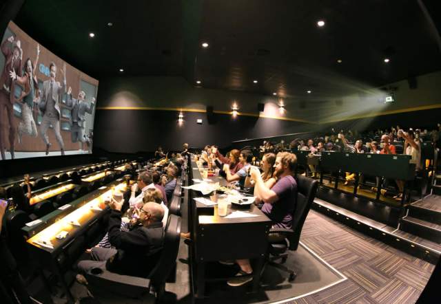 Movie theater auditorium with tiered seating and tables where guests are watching a film on a large screen while eating and drinking, with a projector beam visible and many audience members raising their hands toward the screen.