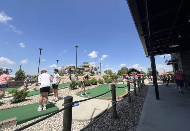 People playing mini golf on an outdoor course under a clear blue sky, with walkways, landscaping, and a building structure along the right side.