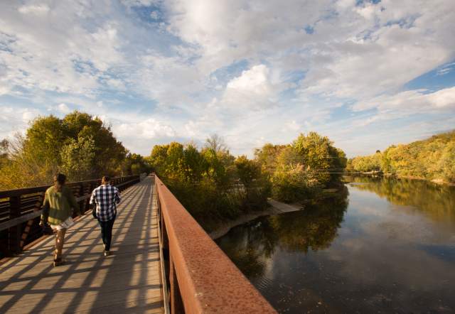 Two people walk along a pedestrian bridge at Forest Park beside a calm river, surrounded by trees under a partly cloudy sky.