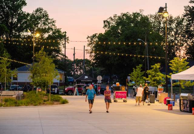 People walk through Grand Junction Plaza at dusk during the June 2023 Jams at the Junction event, with string lights glowing above vendor tents and trees.