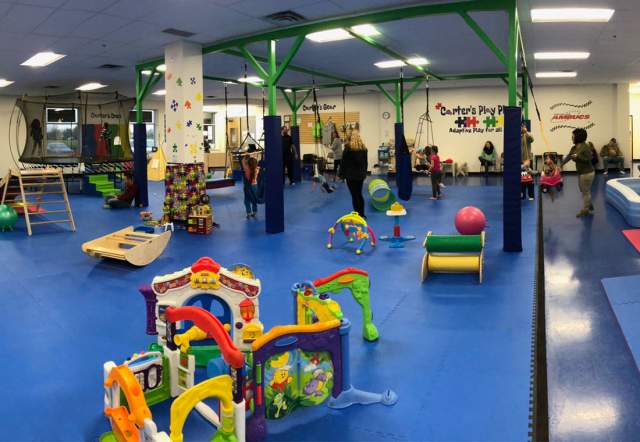 Wide interior view of an indoor play space with blue flooring, colorful soft play equipment, climbing structures, foam blocks, and seating areas, with people scattered throughout the room.