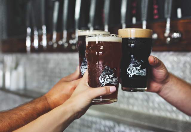 Three hands holding beer glasses raised together in front of a row of tap handles, with the glasses displaying a printed logo and filled with light and dark beer.