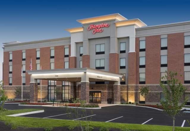 A multi-story hotel with red brick and light-colored accents features a covered entrance and a sign reading “Hampton Inn,” set against a clear evening sky.