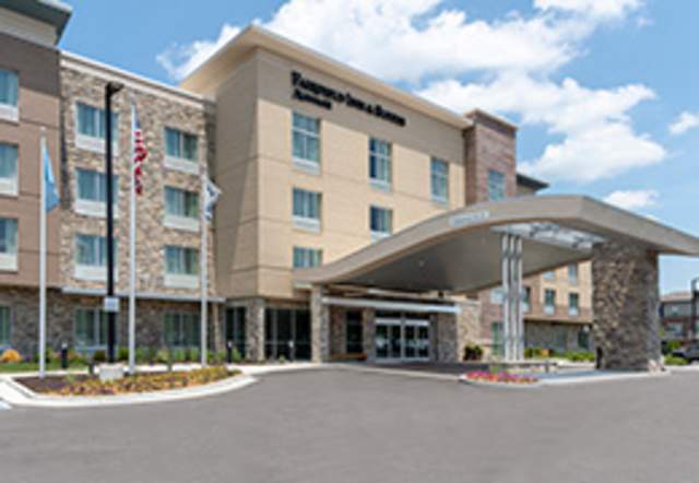 A daytime exterior view of the Fairfield Inn & Suites by Marriott Indianapolis Carmel shows a multi-story hotel building with a mix of tan, stone, and brick façades. A curved covered entrance canopy extends over the driveway leading to the main doors. Large windows line the building, and an American flag stands near the entrance. The hotel sits under a partly cloudy sky, presenting a clean, modern appearance designed for business and leisure travelers.