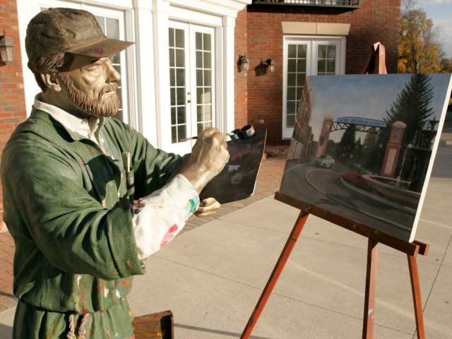 A life-sized statue of a painter captures a scene on canvas, depicting a street with trees and a welcoming archway. The artist stands outdoors near a brick building.