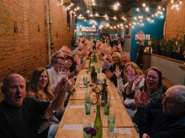 A long group dining table is filled with people seated on both sides, smiling and clapping under warm string lights in a narrow brick-walled patio.