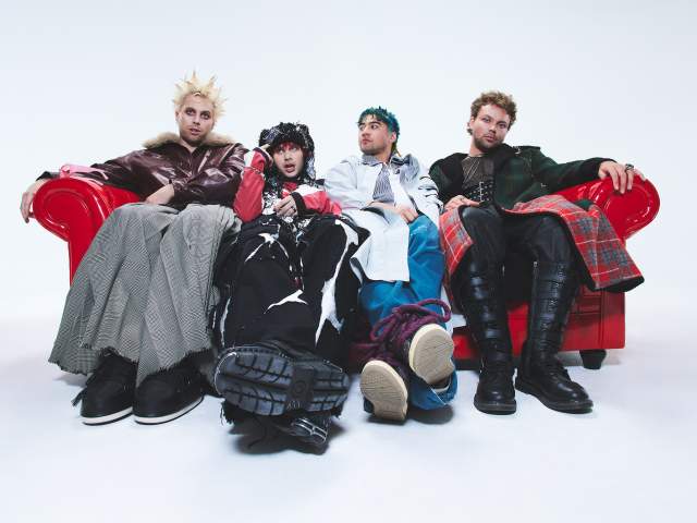 Four people in punk‑inspired outfits sit side by side on a bright red couch against a white background, wearing layered skirts, boots, plaid, and edgy streetwear styles.