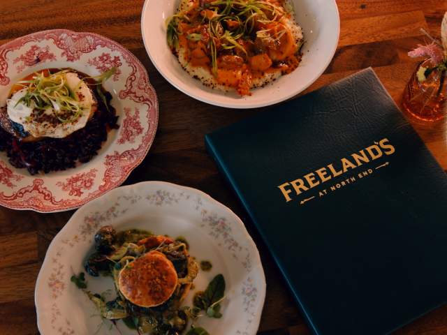Three elegantly plated dishes on a wooden table, featuring a menu titled "Freeland's at North End" and a small floral arrangement.