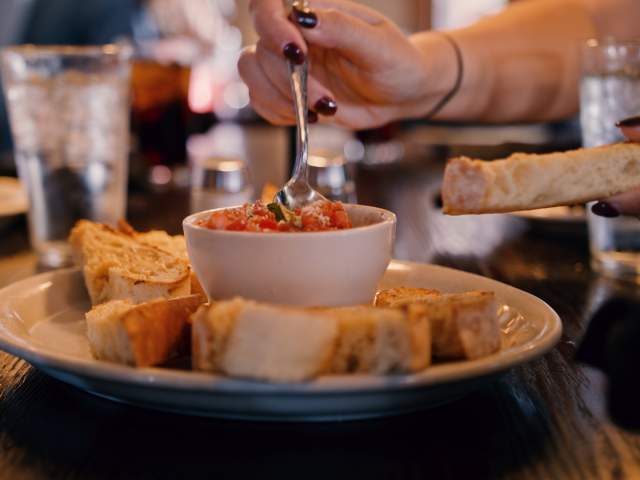 A plate of toasted bread slices surrounds a small bowl of tomato dip. A hand holds a spoon, ready to serve. Glasses of water are in the background.