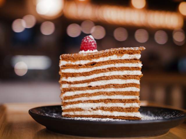 A slice of layered cake with cream and a raspberry on top, presented on a black plate against a softly blurred background.