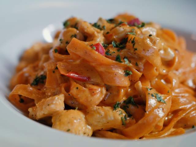 Creamy fettuccine pasta topped with herbs and red peppers, served in a white bowl. The dish is rich and inviting, showcasing a gourmet meal.
