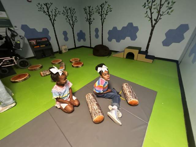 Two children sit on a mat in a nature-themed play area with tree murals, balance logs, and soft play features designed for exploration and sensory engagement.
