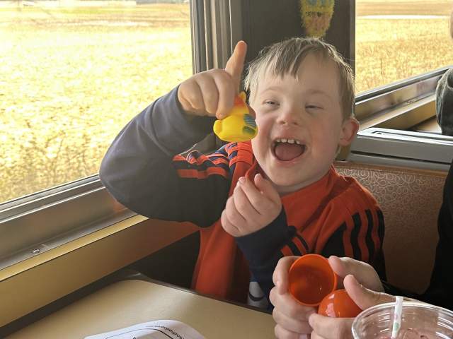 A person seated at a table inside a train car holds up a small yellow toy duck near a window, with coloring pages, crayons, and a drink visible on the table and farmland passing outside.