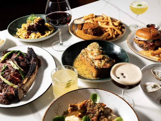 Overhead view of a white table set with an assortment of plated restaurant dishes, including steaks, pasta, a burger with fries, oysters, and elegant cocktails and wine, arranged for a shared dining spread.