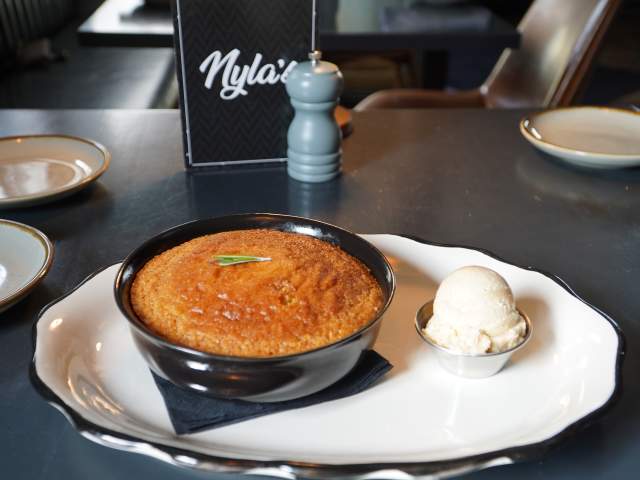 A warm dessert served in a black bowl, accompanied by a scoop of ice cream, on a white plate with a black napkin. A menu card is visible in the background.