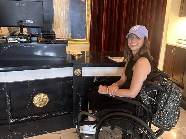 A woman using a wheelchair sits at the lowered, accessible check-in counter inside Hotel Carmichael. She’s wearing a lavender baseball cap and black outfit, smiling toward the camera. Behind the desk are elegant details—black and gold finishes, marble flooring, red velvet curtains, and framed portraits—reflecting the hotel’s classic, upscale decor.