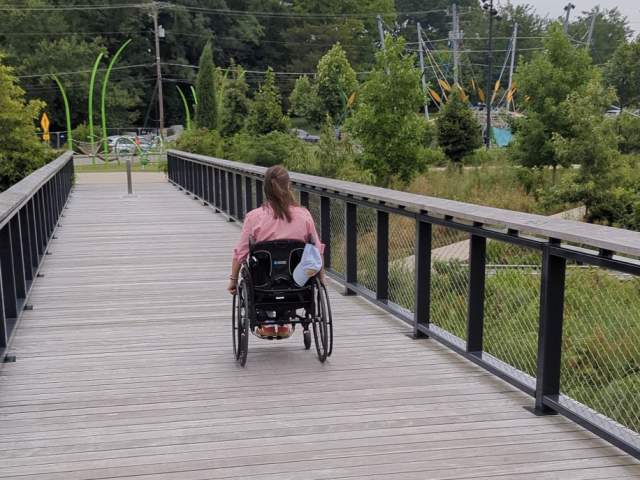 A person using a wheelchair moves across a wooden pedestrian bridge at Grand Junction Plaza, surrounded by greenery.