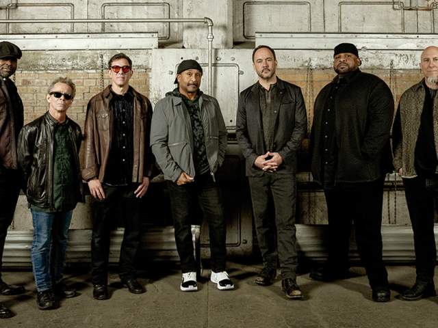 Seven musicians stand side by side in an industrial warehouse setting, wearing dark, casual jackets and jeans against a backdrop of exposed brick, pipes, and metal panels.