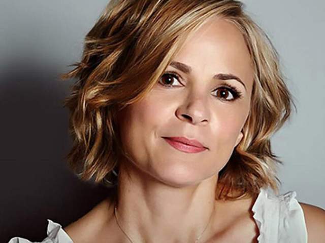 Head-and-shoulders studio portrait of a person with short, wavy hair wearing a light-colored sleeveless top against a gray background.