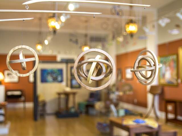 Hanging wooden sculptures in a bright art gallery, with colorful paintings on the walls and warm lighting creating an inviting atmosphere.