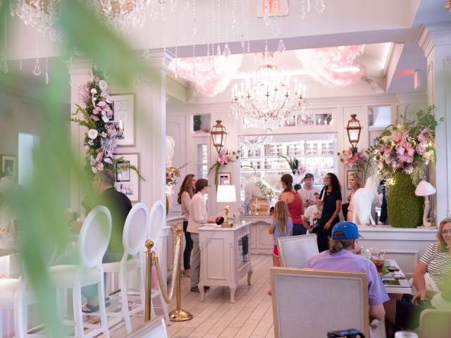 Brightly lit café interior with elegant chandeliers, floral decorations, and patrons engaging at the counter and tables. A welcoming atmosphere.
