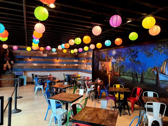 Colorful paper lanterns hang from the ceiling of a vibrant dining space with wooden tables and mixed-color chairs. A mural depicts a serene outdoor scene.