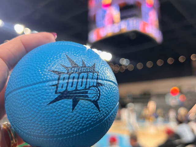 Blue basketball with the Noblesville Boom logo held courtside with a scoreboard blurred in the background.