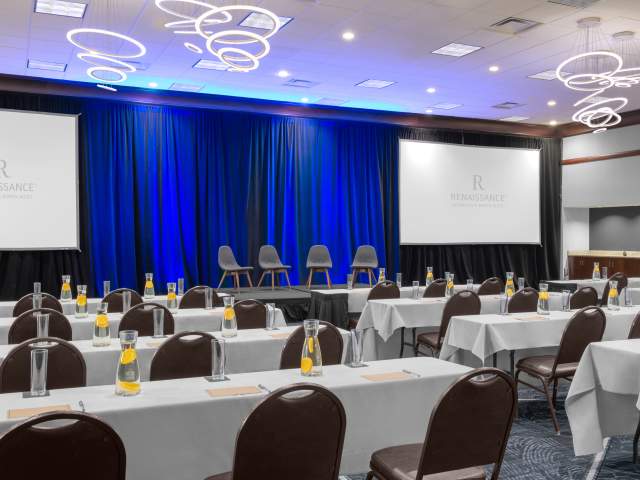 Conference room set in a classroom layout with rows of tables facing a stage with four chairs. Two large projection screens flank blue stage curtains, and modern circular light fixtures hang from the ceiling.