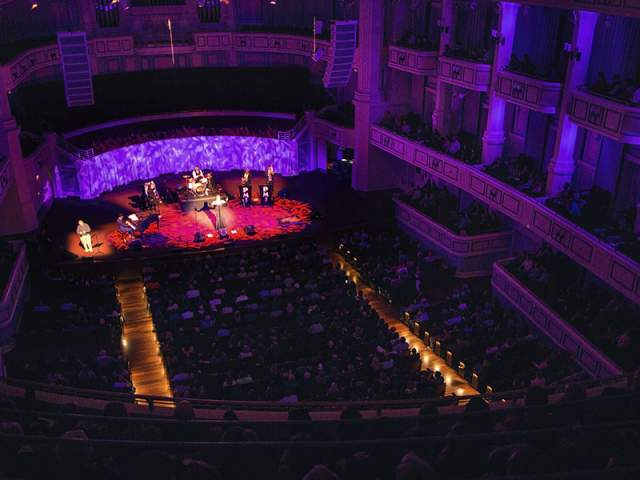 An interior view of a grand concert hall with tiered seating and a stage lit with purple and red lights.