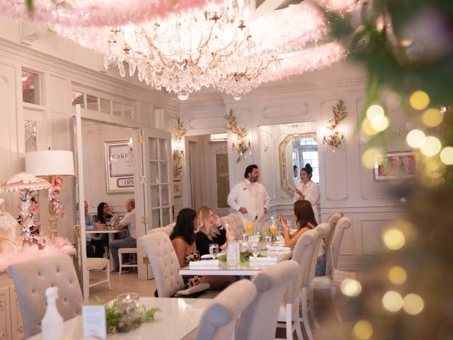 Guests dine in a bright, elegant room at The Cake Bake Shop, decorated with chandeliers, soft pastel accents, and holiday-style lights while servers attend to tables.
