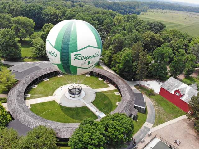 Hot air balloon in vibrant green and white colors floating above a grassy field, trees and a barn below