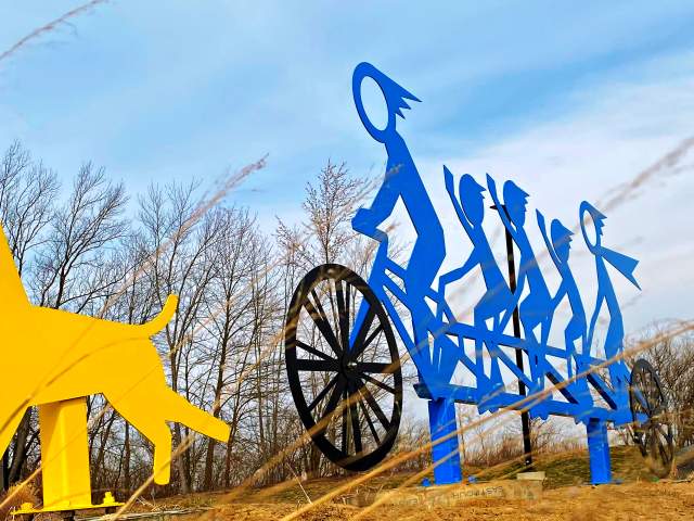 The "Embrace the Trace" outdoor sculpture made of blue, abstract human figures riding a bike and a yellow dog, with a bare tree and a blue sky in the background.