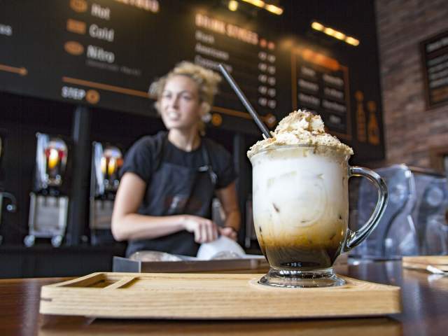 A person behind a coffee bar preparing a large iced coffee topped with foam.