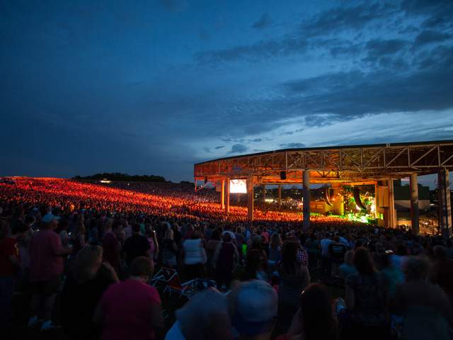 Large outdoor concert venue at dusk, with a brightly lit stage and a crowd filling the seating area. Rows of lights glow across the venue under a darkening blue sky.