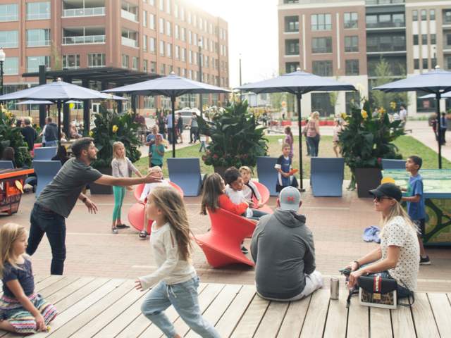 A lively urban street with people walking, children playing, outdoor dining, and brick buildings