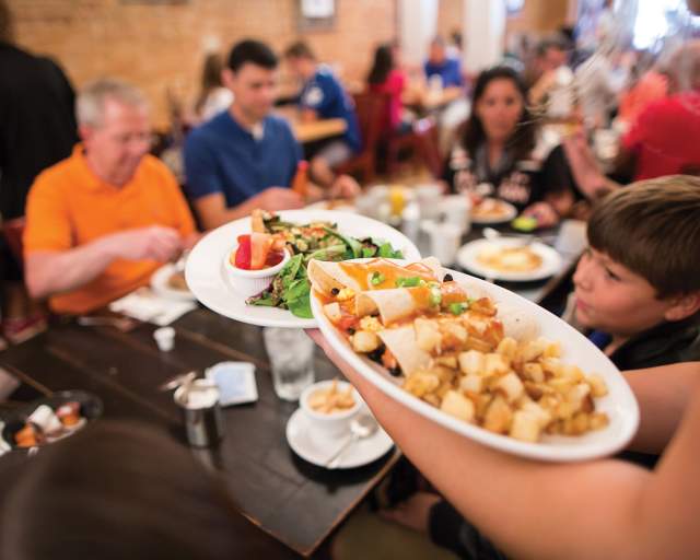 A server carries two large plates of food toward a crowded dining table. Several people of different ages sit around the table with plates, drinks, and utensils, in a busy indoor restaurant setting with warm lighting and brick walls.