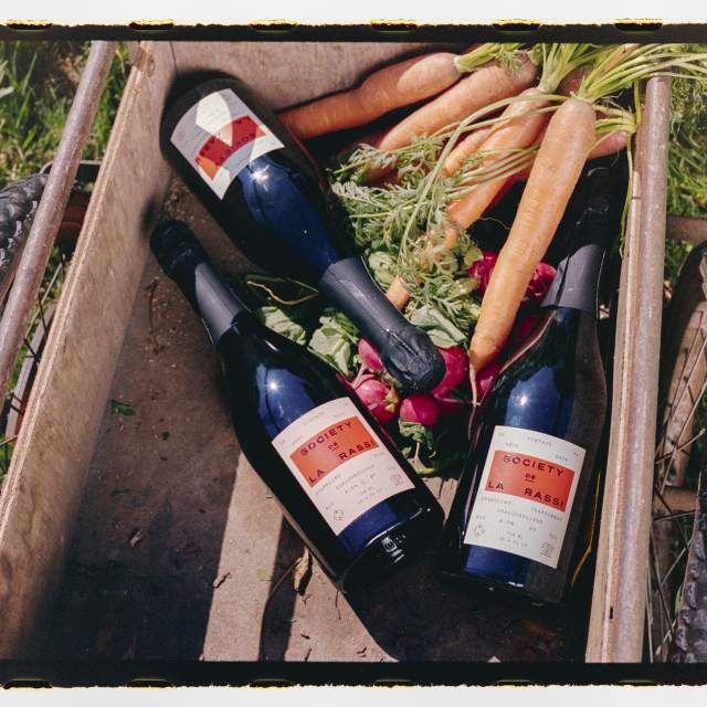 Top-down view of a rustic wooden garden cart holding three bottles of Society de La Rassi sparkling wine alongside freshly harvested carrots with greens and red radishes, set on grass.