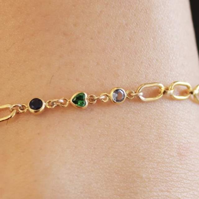Close-up of a gold chain bracelet on a wrist, featuring small bezel-set stones including a black round gem, a green heart-shaped gem, and a clear round gem.