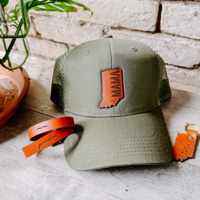 Olive green trucker hat with an orange Indiana-shaped patch reading “MAMA,” displayed on a tabletop beside an orange leather keychain and stacked leather bracelets, with a potted plant and white brick wall in the background.