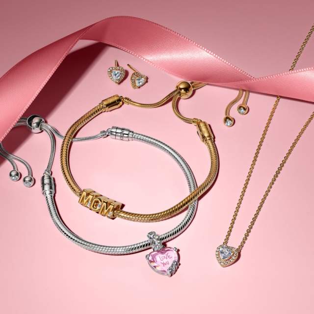Gold and silver charm bracelets with heart-shaped charms and messages, paired with heart stud earrings and a heart pendant necklace, arranged on a pink background with a satin ribbon.