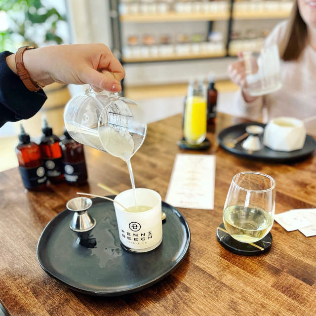 Hand pours melted candle wax from a glass measuring cup into a white Benn & Beech candle jar set on a wooden table, with fragrance bottles, tools, and drinks visible in a candle-making workshop setting.