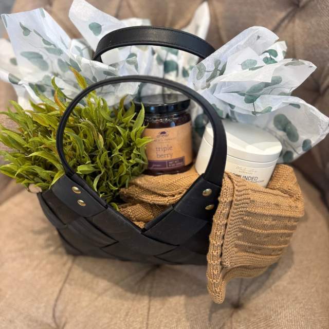 Black woven gift basket holding a jar of berry jam, a white candle, green foliage, and a folded knit item, wrapped with patterned tissue paper and placed on a cushioned chair.
