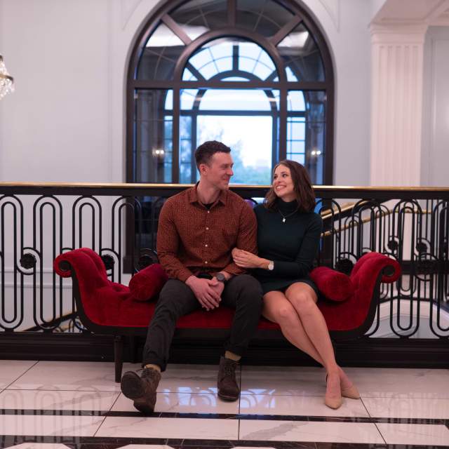 A couple sits together on a red velvet bench inside Feinstein’s at Hotel Carmichael, smiling at each other in an elegant lobby with chandeliers and ornate railings.